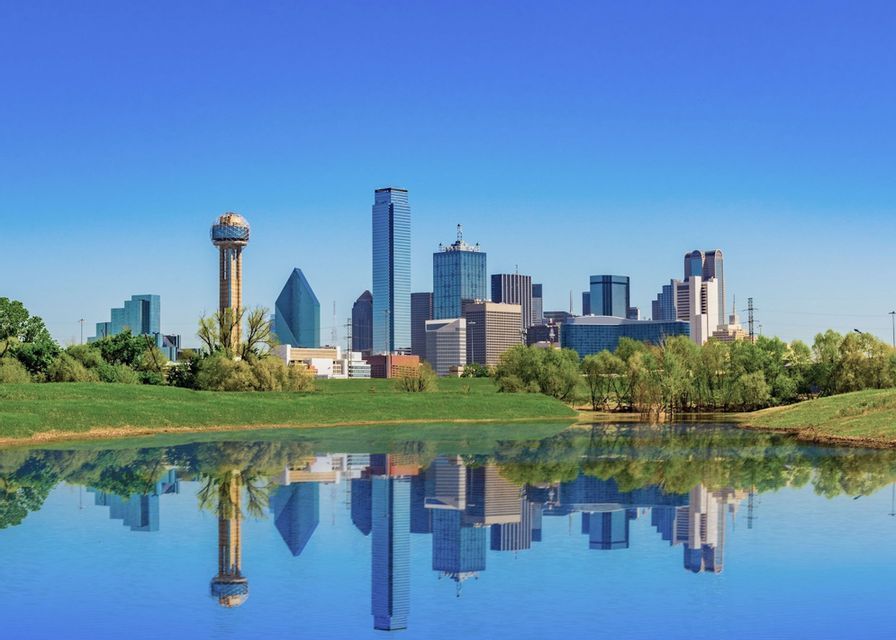 A modern city skyline with skyscrapers reflected perfectly in calm water, with a green grassy bank in the foreground.