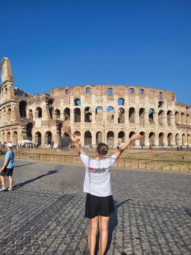 Ein WeRoad-Reiseleiter mit ausgestreckten Armen, von hinten gesehen, vor dem Kolosseum an einem sonnigen Tag.
