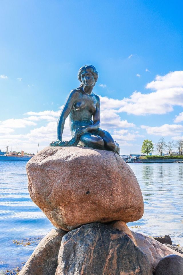 Eine bronzene Meerjungfrauenstatue sitzt auf einem großen Felsen am Meer unter blauem Himmel mit weißen Wolken.