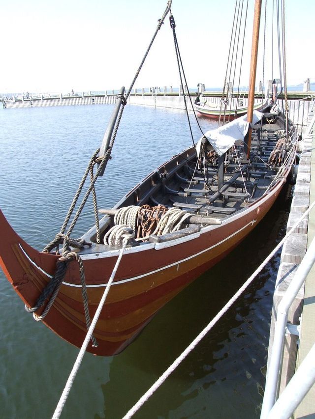 Eine hölzerne Wikinger-Langschiff-Replik mit aufgewickelten Seilen und einem Mast liegt an einem Holzsteg vertäut.