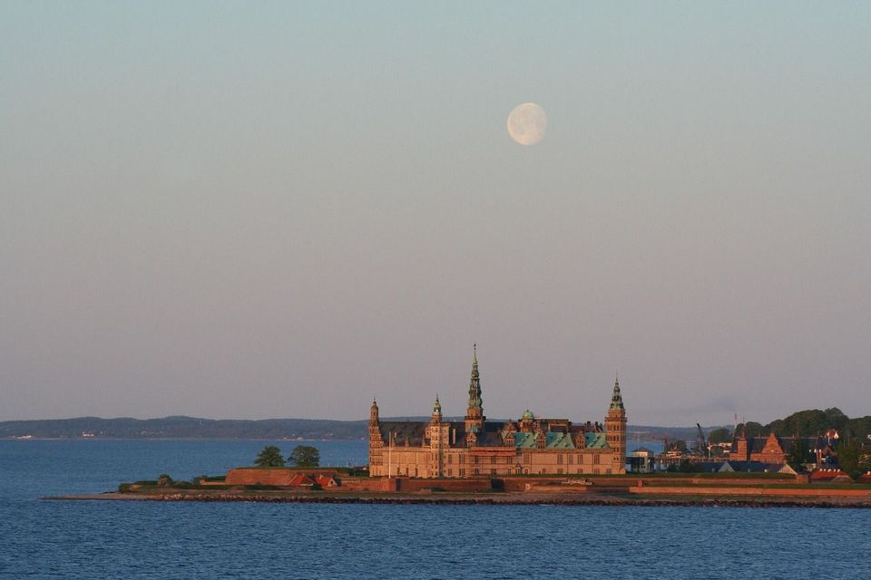 Ein großes Schloss mit spitzen Türmen thront auf einer Halbinsel am Meer, wobei ein Vollmond am Dämmerungshimmel sichtbar ist.