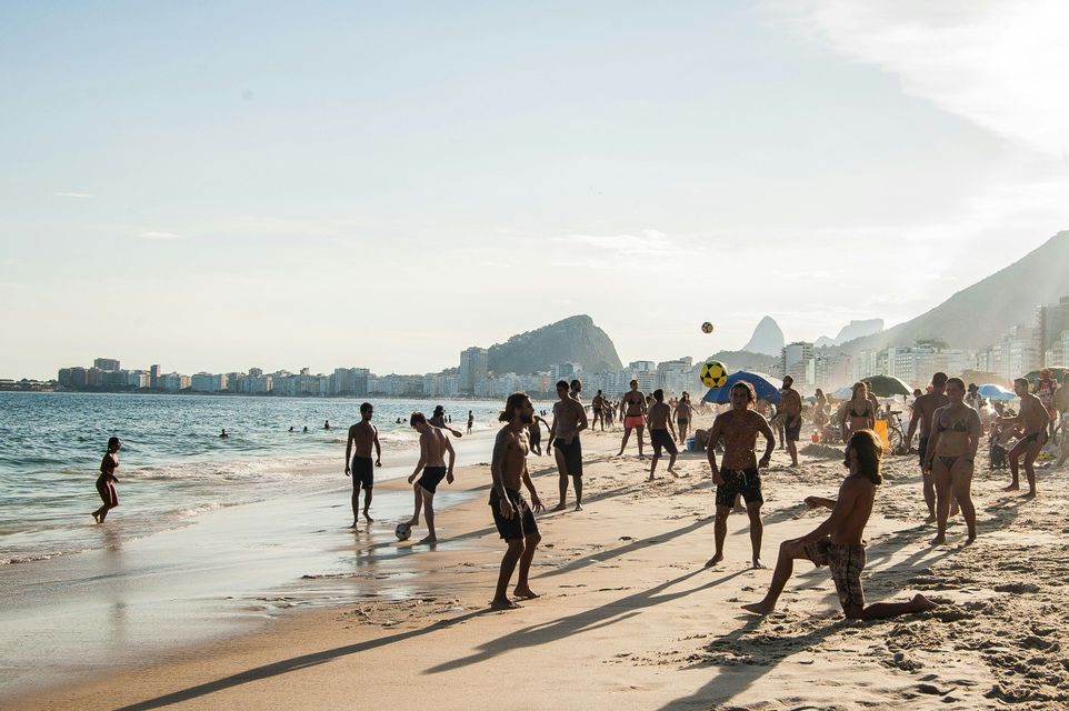 Un gruppo di persone gioca a pallone su una spiaggia sabbiosa, con lo skyline di una città e montagne sullo sfondo.