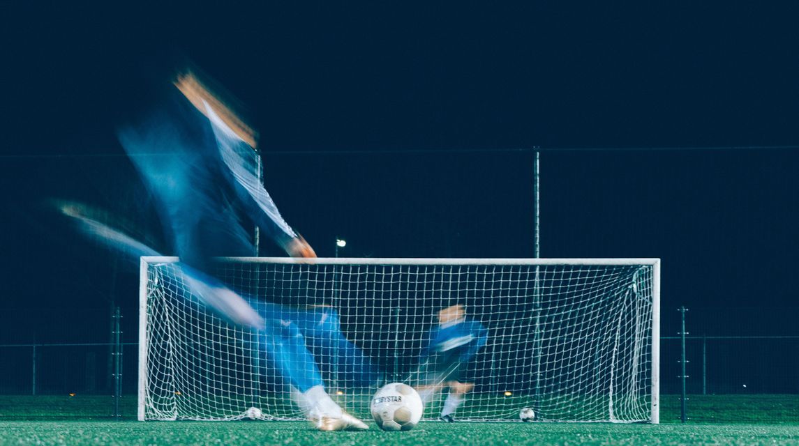 Una vista sfocata di un calciatore che calcia un pallone verso una porta difesa da un portiere di notte.