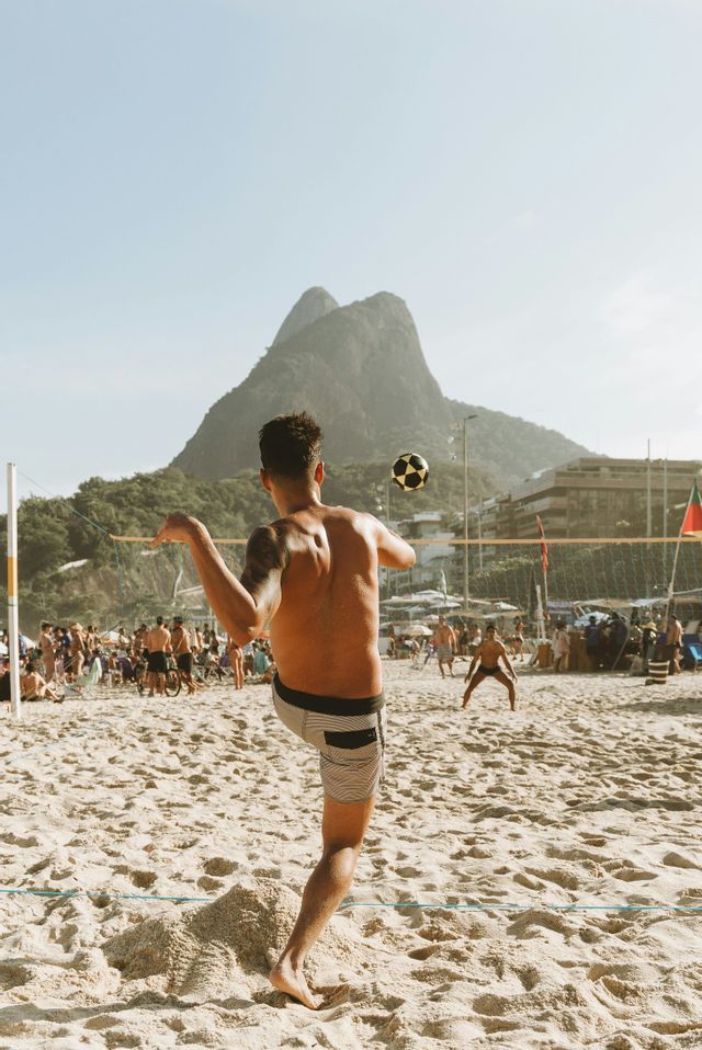 Un uomo ripreso di spalle calcia un pallone da calcio in aria durante una partita su una spiaggia affollata con una montagna sullo sfondo.