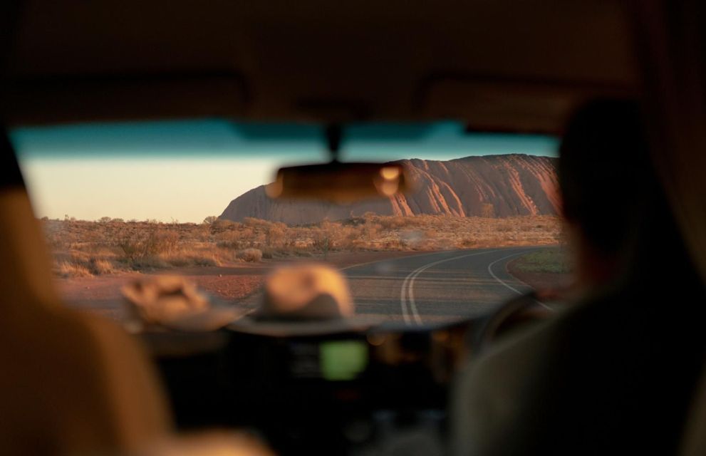 Blick aus dem Auto: Eine WeRoad-Gruppenreise fährt auf einer kurvenreichen Straße einer großen roten Felsformation bei Sonnenuntergang entgegen.