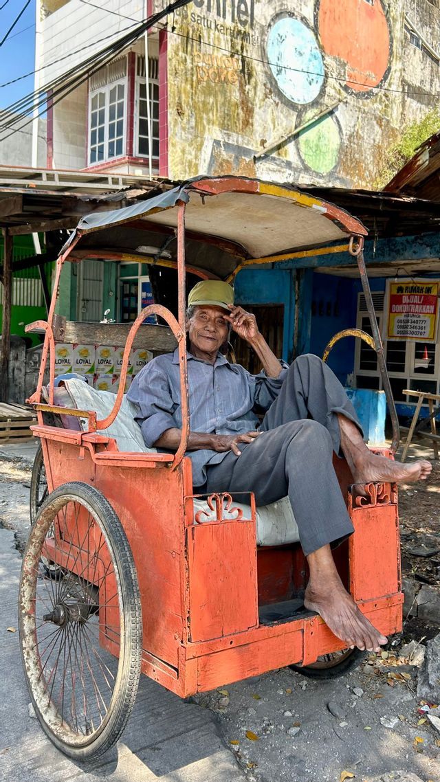 A smiling elderly man with a hat sits barefoot in a traditional orange three-wheeled rickshaw on a sunny street.