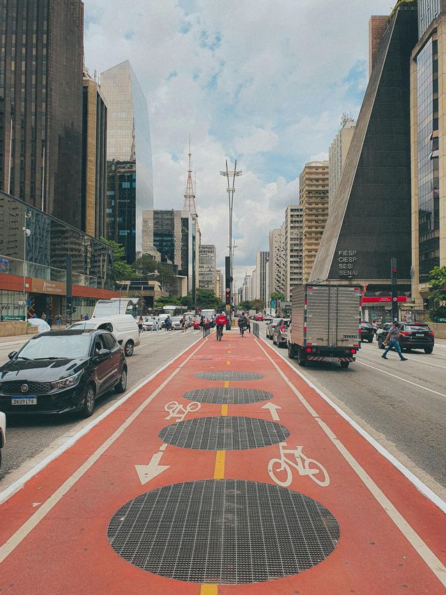 Un carril bici ancho y rojo con símbolos de bicicleta recorre el centro de una ajetreada calle urbana flanqueada por rascacielos.