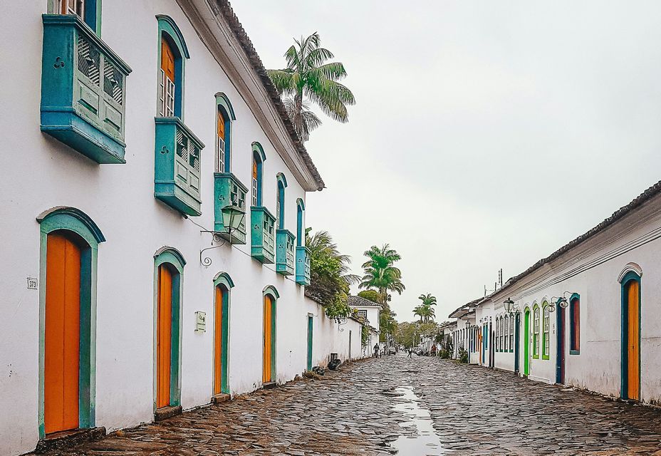 Una calle adoquinada mojada flanqueada por edificios coloniales encalados con puertas, ventanas y balcones coloridos bajo un cielo cubierto.