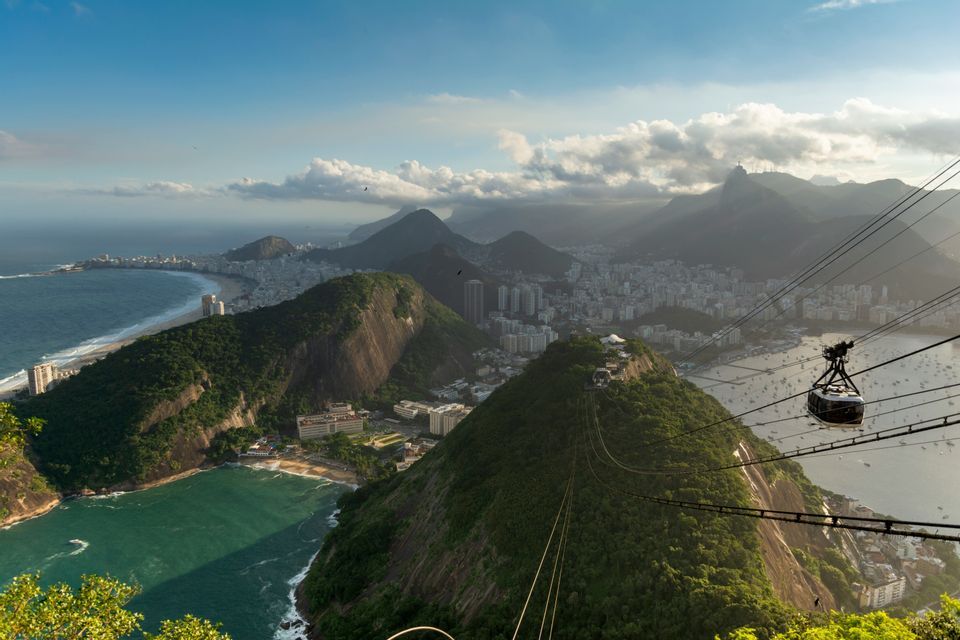 Un teleférico se mueve a lo largo de sus cables sobre una exuberante montaña verde, con vistas a una extensa ciudad costera y una bahía con barcos.