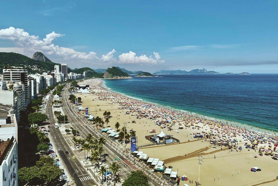 Una vista aérea de una playa concurrida con sombrillas de colores junto a un horizonte urbano y el océano azul bajo un cielo parcialmente nublado.