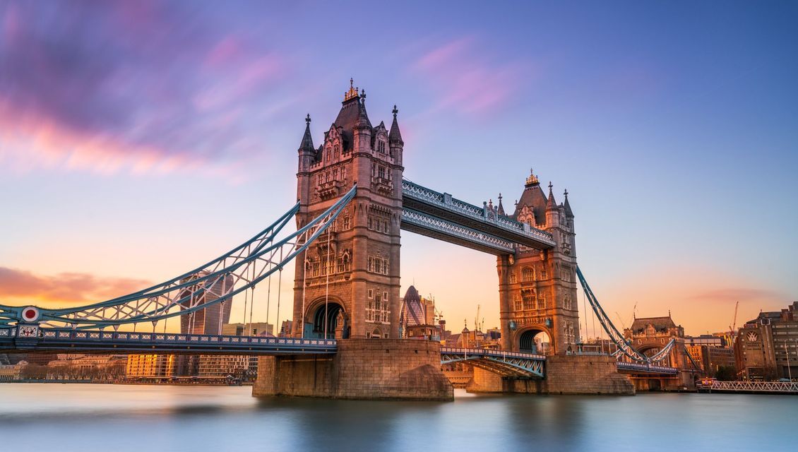 A long-exposure shot of the Tower Bridge in London spanning the River Thames at sunset, under a purple and orange sky.