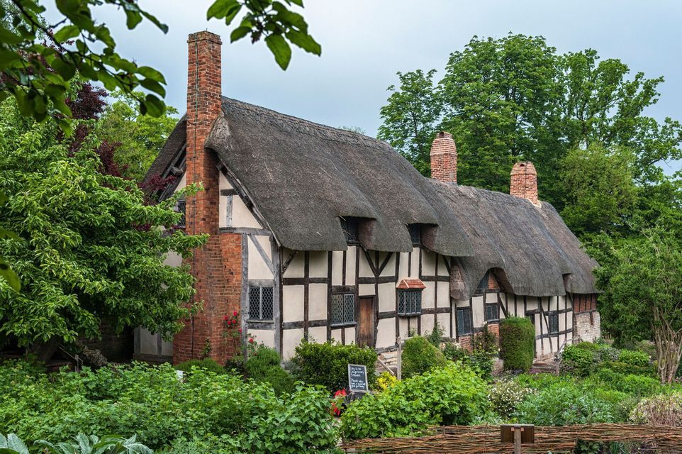 A Tudor-style timber-framed house with a thatched roof and brick chimneys, surrounded by a lush green garden.