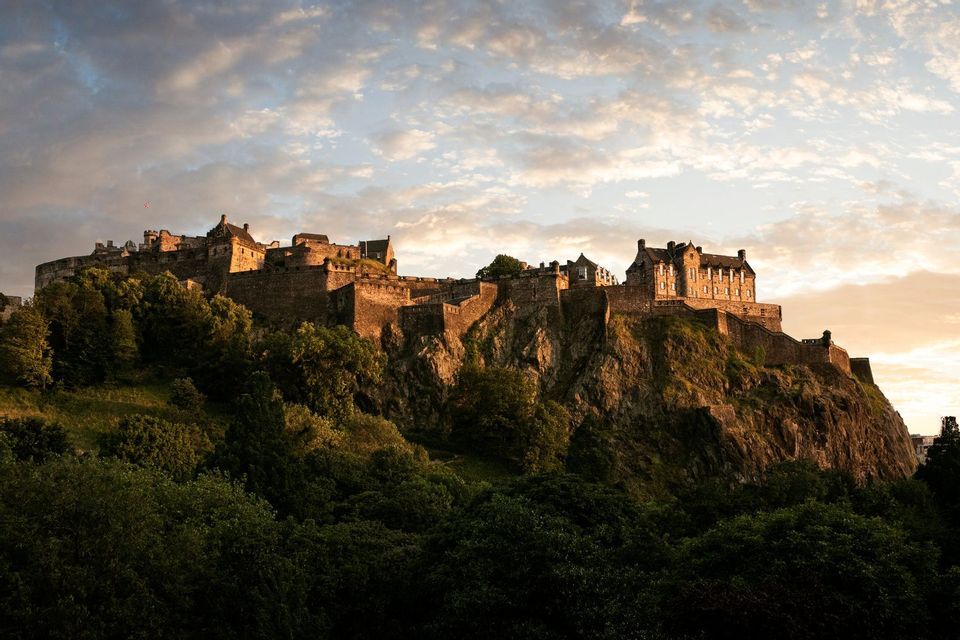 A large stone castle sits atop a rocky, tree-covered hill, illuminated by sunset against a cloudy sky.