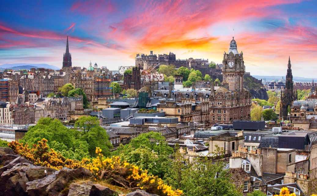 A historic European cityscape featuring a castle and clock towers, viewed from a hill with yellow flowers under a colorful sunset sky.