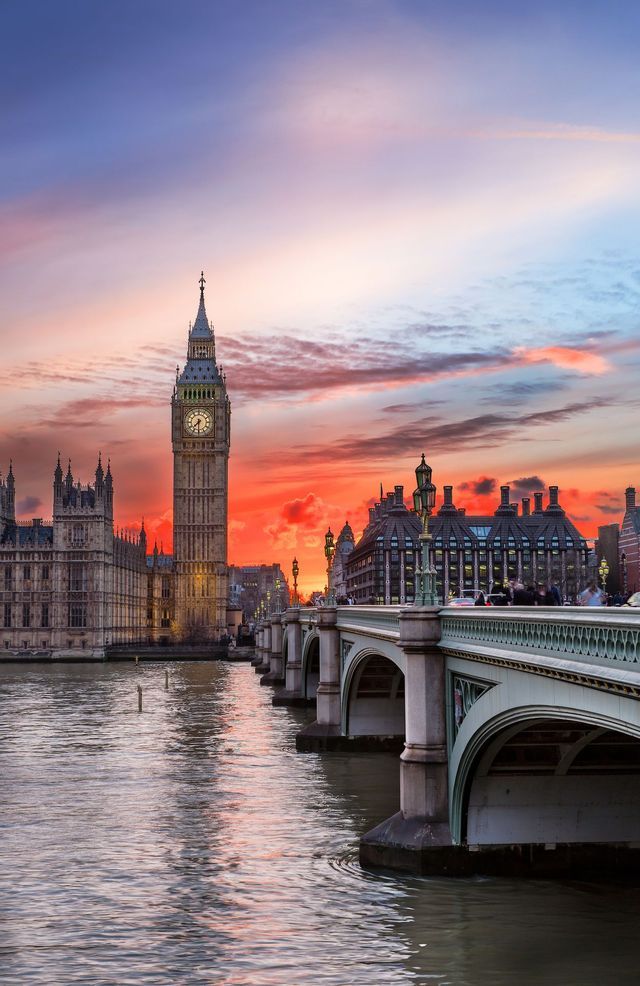A prominent clock tower and government building are reflected in a river at sunset, with a stone bridge in the foreground.