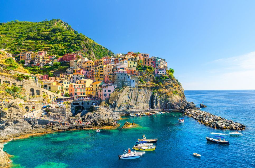A colorful village built on a rocky, terraced cliffside overlooking a bright blue sea with several small boats in a cove.