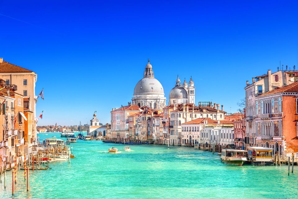 A wide canal with turquoise water, lined with historic buildings and a domed basilica in the distance under a clear blue sky.