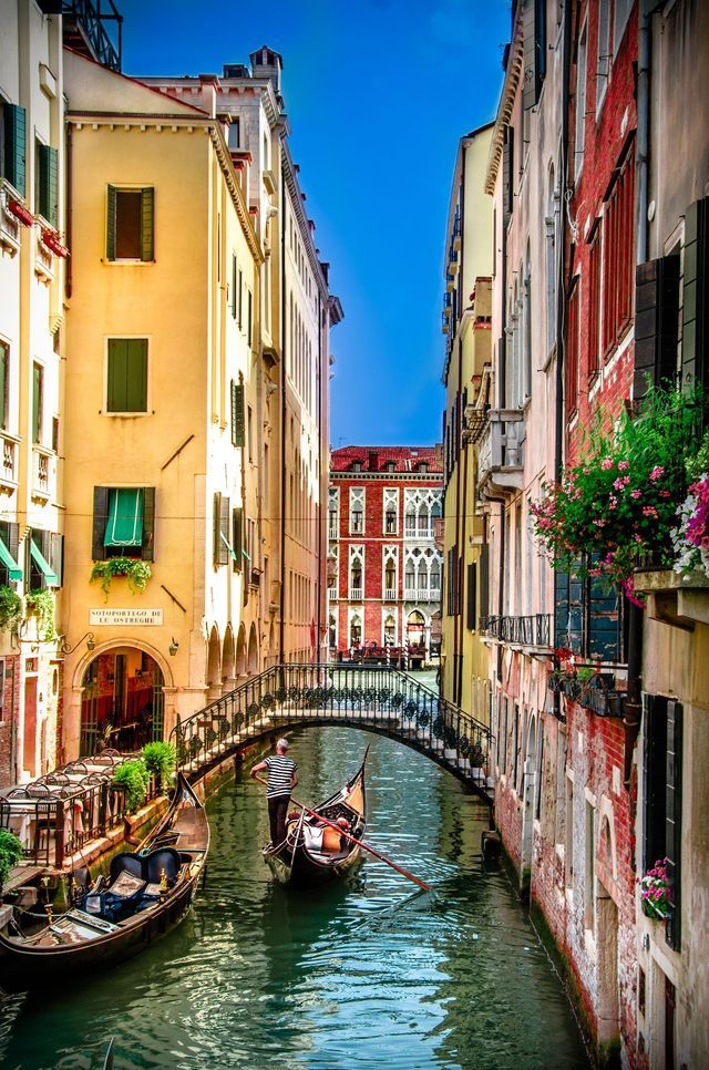 A gondolier in a striped shirt steers a gondola down a canal, passing under an arched bridge between colorful buildings.