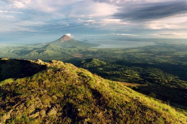 Una vista panoramica da una collina erbosa che domina una valle lussureggiante con un vulcano e un grande lago in lontananza.