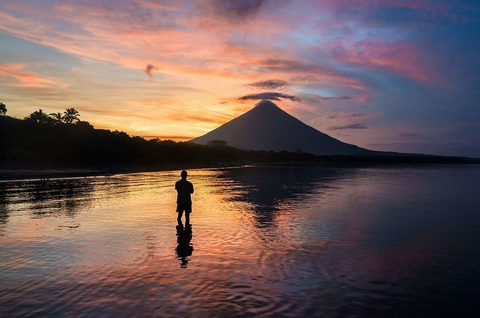 Una silhouette si staglia in acque calme di fronte a un grande vulcano durante un tramonto colorato.