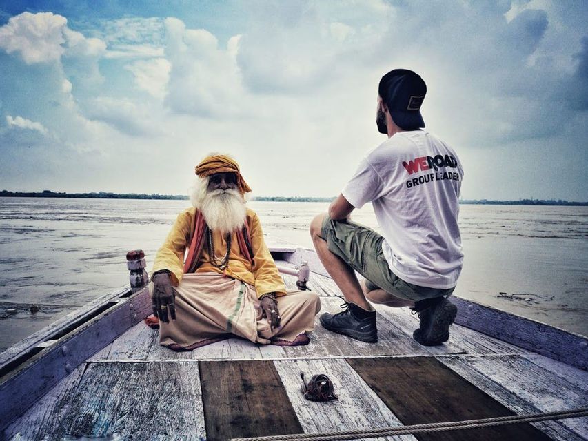 Un capogruppo WeRoad siede con un anziano signore con una lunga barba bianca a prua di una barca di legno su un fiume.