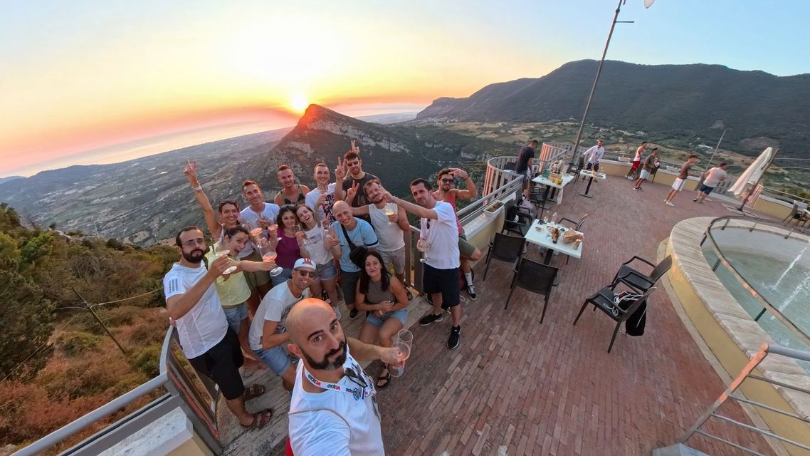 Un viaje en grupo de WeRoad se toma una selfie de gran angular en un mirador panorámico, brindando con bebidas mientras el sol se pone detrás de las montañas.