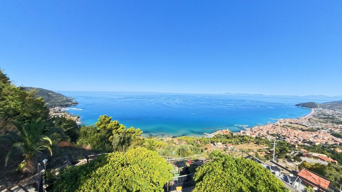 Una vista panorámica desde un punto elevado de un pueblo costero que se curva alrededor de una amplia bahía con agua turquesa bajo un cielo azul claro.