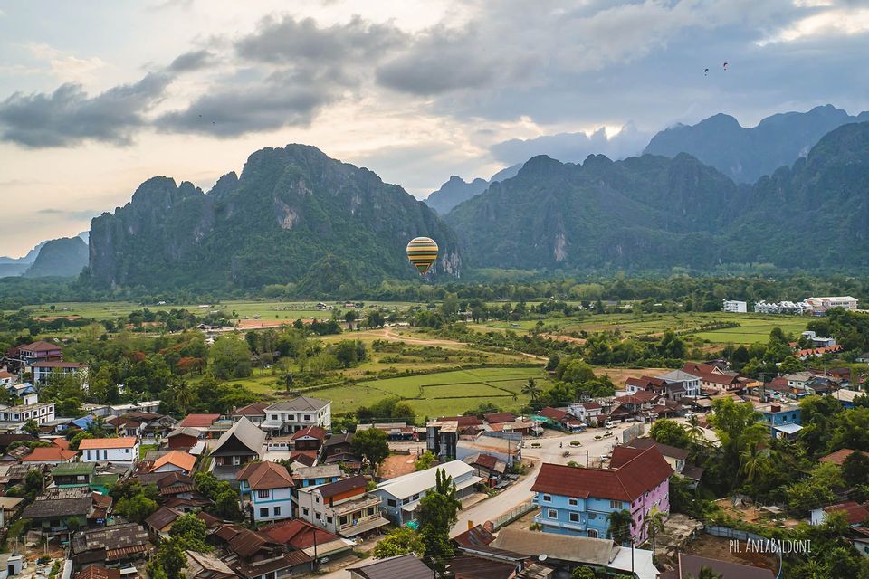Un globo aerostático vuela sobre un pueblo anidado en la base de grandes montañas verdes bajo un cielo nublado.