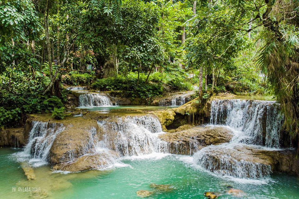 Una cascada escalonada se precipita sobre salientes de piedra en pozas turquesas dentro de un denso bosque verde, donde una persona está nadando.
