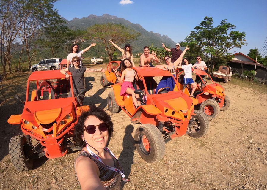 Un viaje en grupo de WeRoad posando para una foto con buggies de dunas de color naranja brillante en un camino de tierra con montañas al fondo.