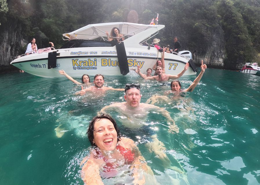 Un grupo WeRoad se toma un selfie nadando en aguas turquesas junto a una lancha rápida cerca de acantilados rocosos.