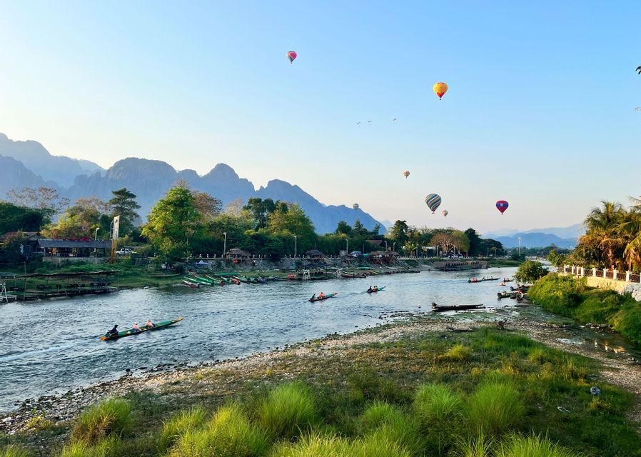 Globos aerostáticos flotan sobre un río con botes de cola larga, con un telón de fondo de montañas bajo un cielo despejado.