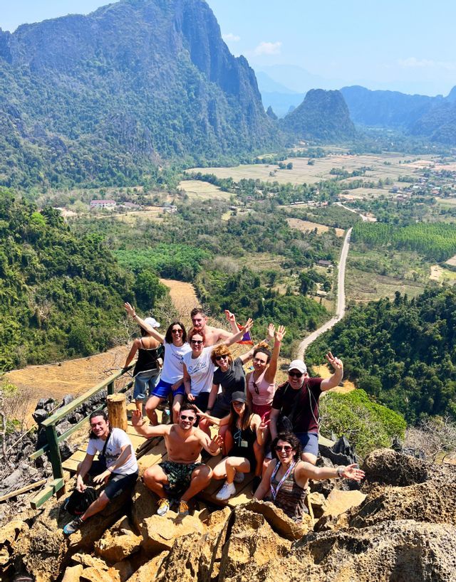 Un viaje en grupo de WeRoad posando juntos en un mirador rocoso en la cima de una montaña, con vistas a un exuberante valle verde y formaciones kársticas.