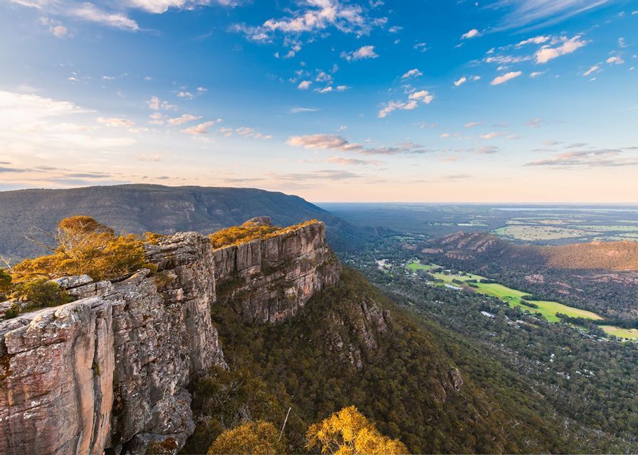 Ein Panoramablick von einer felsigen Klippe, der ein bewaldetes Tal und weite Ebenen unter einem blauen Himmel mit Wolken bei Sonnenuntergang zeigt.