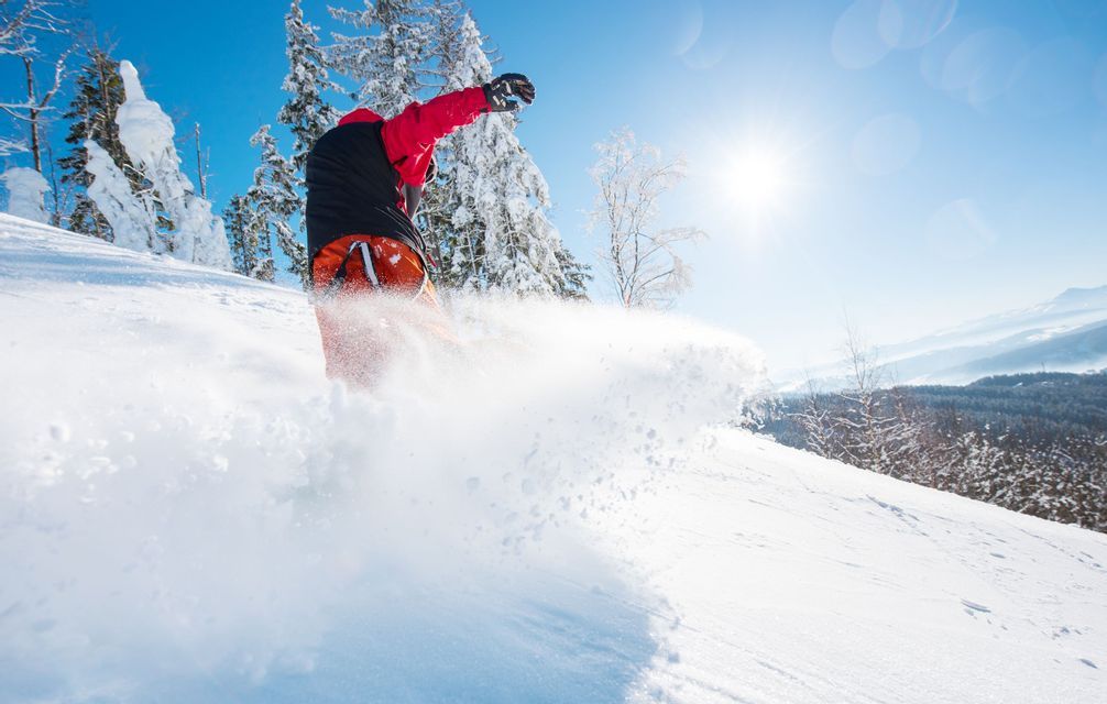 Ein Snowboarder in einer roten Jacke zieht Spuren durch tiefen Pulverschnee auf einem sonnigen Berghang.