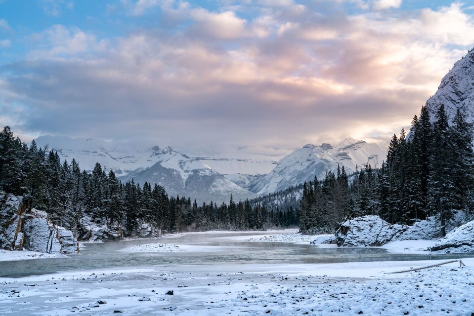 Ein teilweise zugefrorener Fluss fließt durch einen schneebedeckten Kiefernwald mit schneebedeckten Bergen im Hintergrund unter einem bewölkten Himmel.