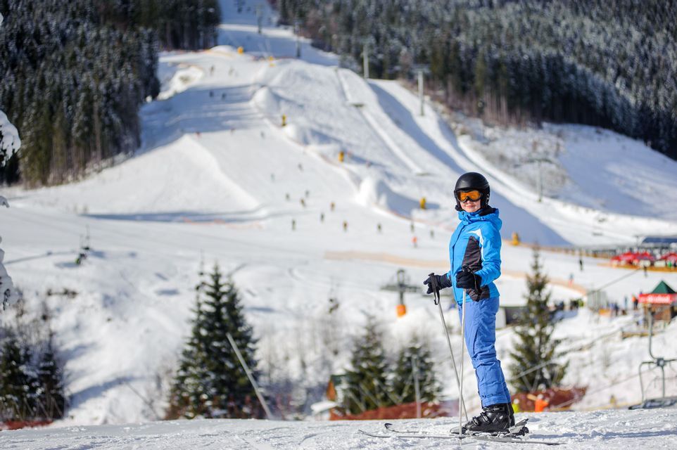 Ein Skifahrer in blauer Jacke und Helm steht auf einem verschneiten Hang in einem Skigebiet mit Kiefern.