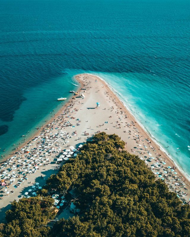 Luftaufnahme eines dreieckigen Sandstrandes, überfüllt mit Menschen und Sonnenschirmen, der sich ins türkisfarbene Meer erstreckt.