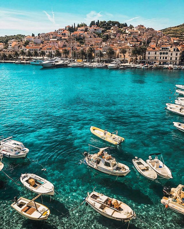 Blick von oben auf kleine Boote, die im klaren türkisfarbenen Wasser ankern, mit einer Küstenstadt auf einem Hügel im Hintergrund.