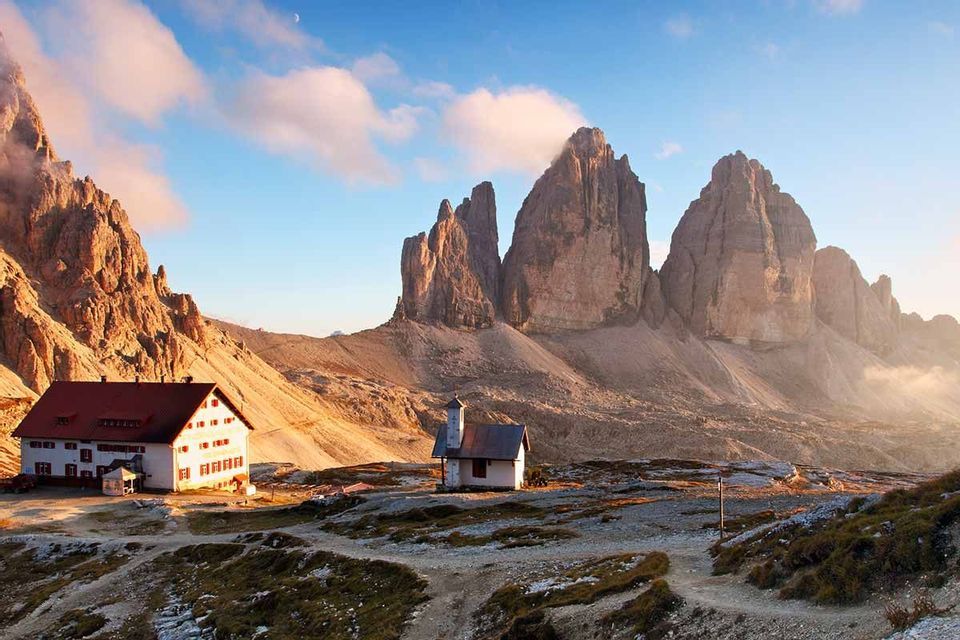 Un rifugio alpino e una piccola cappella si trovano su un altopiano roccioso ai piedi di cime frastagliate sotto un cielo azzurro con una luna crescente.