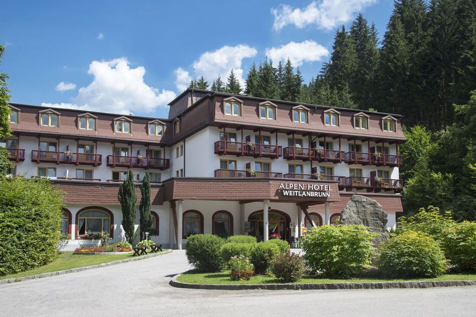 L'esterno dell'Alpen Hotel Weitlanbrunn, circondato da giardini paesaggistici e una fitta pineta sotto un cielo parzialmente nuvoloso.
