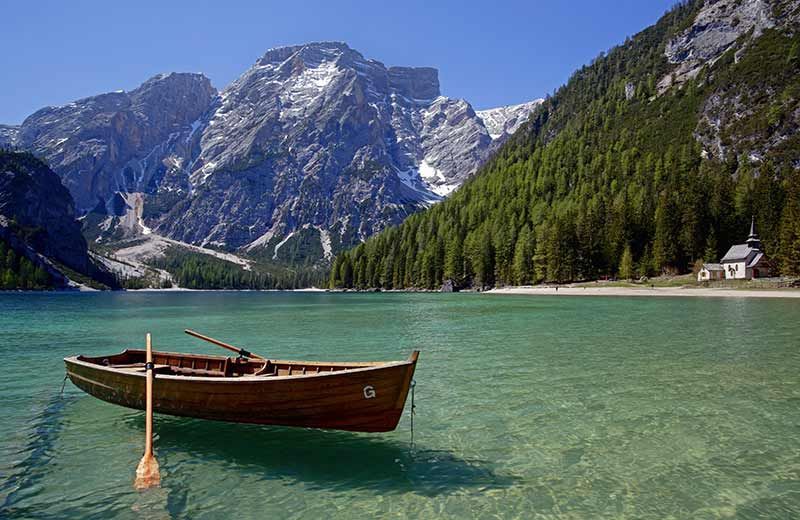 Una barca a remi di legno galleggia su un lago alpino cristallino, con montagne innevate e una pineta sotto un cielo blu.