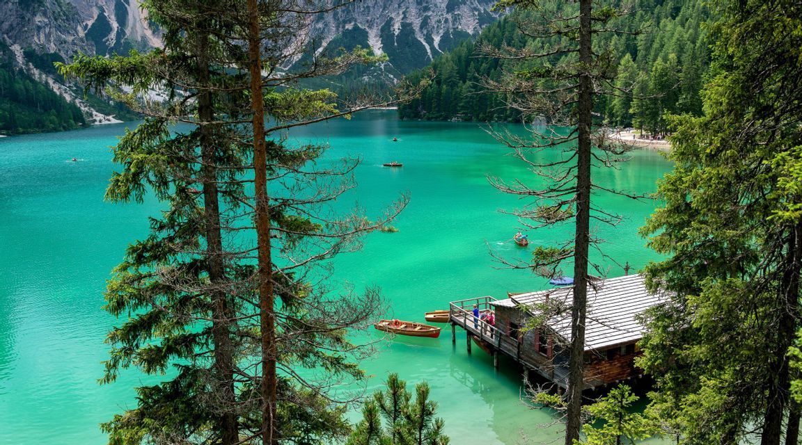 Una vista tra due pini di un lago alpino turchese con una rimessa per barche in legno, barche a remi e montagne lontane