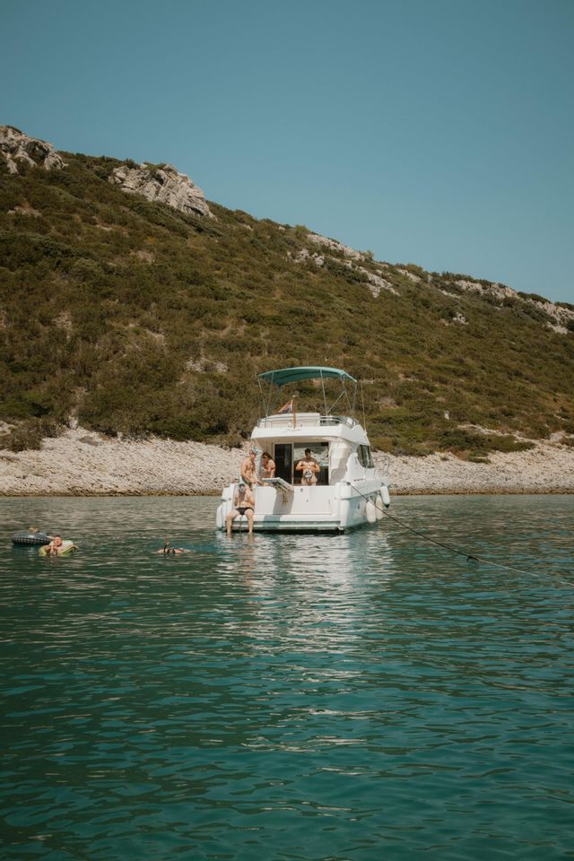 Eine WeRoad-Gruppenreise entspannt auf einer weißen Yacht, die in einem ruhigen Meer ankert, mit einigen Leuten, die neben einer grünen, felsigen Küste schwimmen.