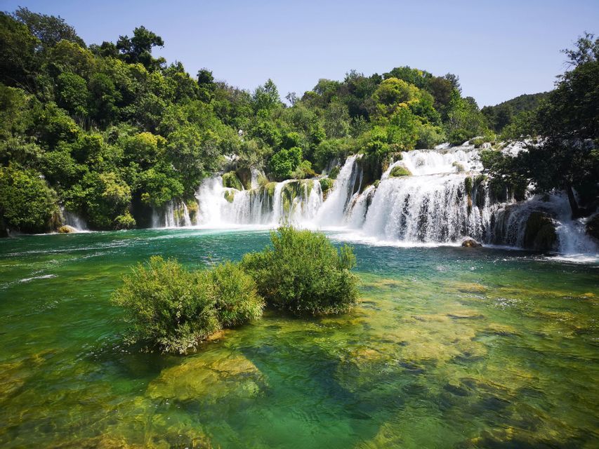 Mehrere breite Wasserfälle stürzen in einen klaren, türkisen Fluss, umgeben von einem dichten grünen Wald unter klarem Himmel.