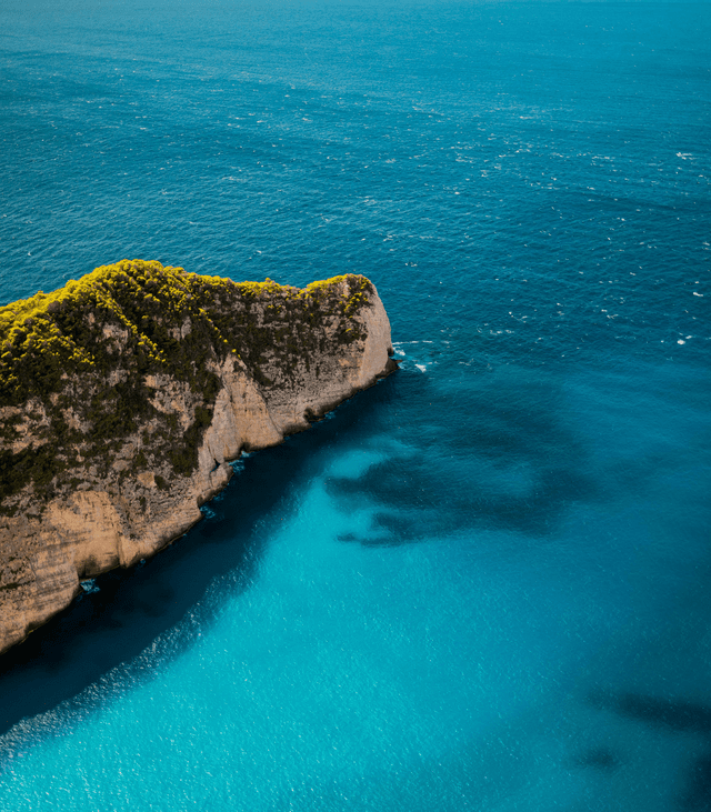 Luftaufnahme einer felsigen, grün bewachsenen Klippe, die in ein türkisfarbenes und tiefblaues Meer ragt.