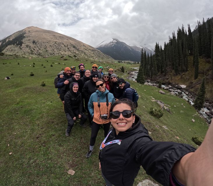A WeRoad group trip poses for a selfie on a grassy mountainside, with a river and snow-capped peaks in the background.