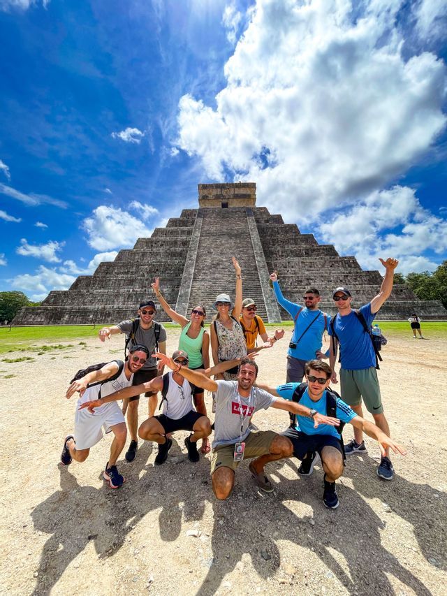 A WeRoad group trip of friends posing for a photo in front of a large stone step pyramid under a bright blue sky.