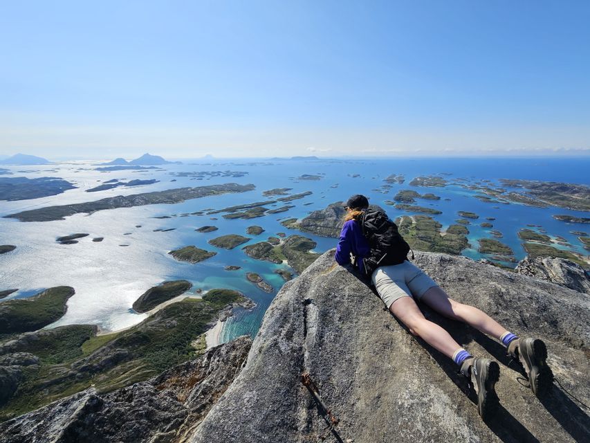 A hiker with a backpack lies on a rocky summit, looking out over a vast archipelago with turquoise water under a clear sky.