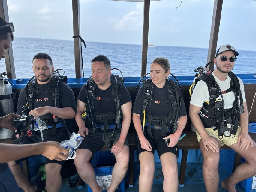 Un groupe WeRoad de quatre personnes en équipement de plongée complet, assises sur un bateau, se préparant à plonger en pleine mer.